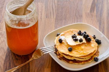 Top angle view on white plate on the wooden board table with pancakes and butter blueberries over and maple syrup or honey with fork and knife by the plate