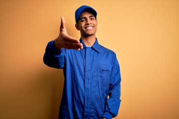 Young african american mechanic man wearing blue uniform and cap over yellow background smiling friendly offering handshake as greeting and welcoming. Successful business. © Krakenimages.com