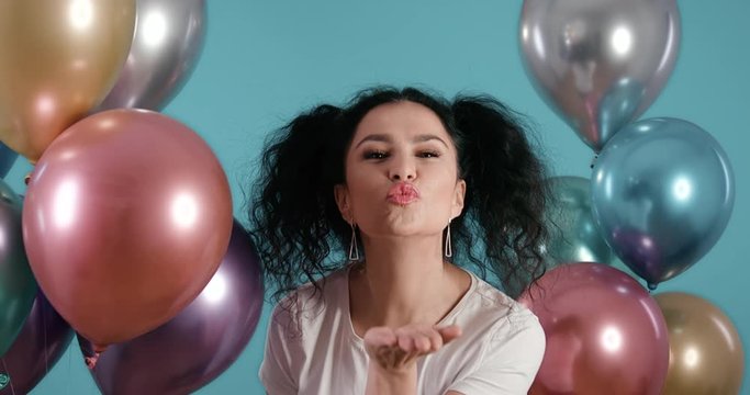 Portrait of beautiful young woman with black curly hair smiling and kissing towards the camera with ballons in her hands and blue background shot in 4k super slow motion