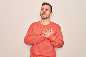 Young handsome man with blue eyes wearing casual sweater standing over white background smiling with hands on chest with closed eyes and grateful gesture on face. Health concept.