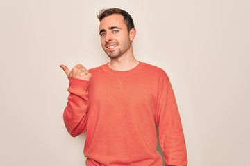 Young handsome man with blue eyes wearing casual sweater standing over white background smiling with happy face looking and pointing to the side with thumb up.