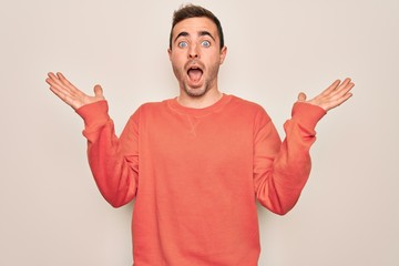 Young handsome man with blue eyes wearing casual sweater standing over white background celebrating crazy and amazed for success with arms raised and open eyes screaming excited. Winner concept