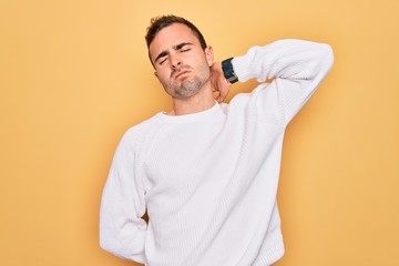 Young handsome man with blue eyes wearing casual sweater standing over yellow background Suffering of neck ache injury, touching neck with hand, muscular pain
