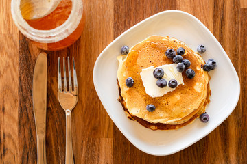 Top angle view on white plate on the wooden board table with pancakes and butter blueberries over and maple syrup or honey with fork and knife by the plate