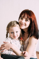 Blurred Portrait of mom and daughter on a white background. Mom brunette holds her blonde daughter in her arms while sitting on the couch. Mother's care for the baby. Mother's day concept.