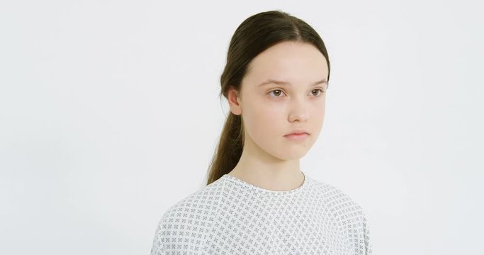 Camera moving round sad unhealthy young Caucasian girl in hospital night dress on white wall background. Portrait of tired upset sick female medical patient with bad look. Close up of face.