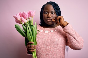 Young african american plus size woman with braids holding bouquet of pink tulips flower mouth and...