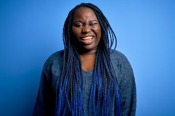 African american plus size woman with braids wearing casual sweater over blue background winking looking at the camera with sexy expression, cheerful and happy face.