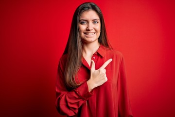 Young beautiful woman with blue eyes wearing casual shirt standing over red background cheerful with a smile of face pointing with hand and finger up to the side with happy and natural expression