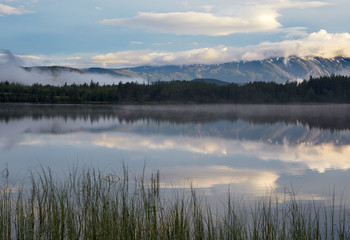 Morning. Dawn. Sky and clouds are reflected on the surface of the water. In the distance, the opposite shore of the lake and a mountain range are visible.