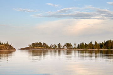 Obraz premium Summer. Evening.Calm water surface of the lake. In the distance you can see a narrow strip of rocky shore,overgrown with moss and trees.On the left is the passage between the Islands. Gentle blue sky.