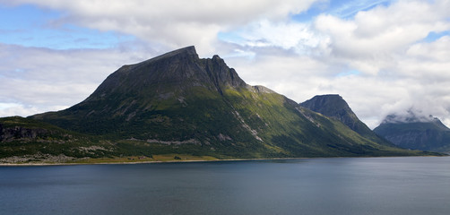 Norway. Summer. In the foreground is a bay with dark blue water. In the background are the rocky peaks of the fjord. Green grass and shrubs cover the mountain range. White, fluffy clouds and blue sky.