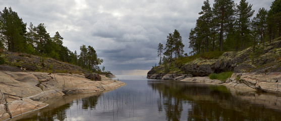 Summer. In the foreground we see the Bay of lake Ladoga, surrounded on the left and right by rocky shores. The upper part of the banks is covered with pine trees.Quiet, calm water. A dark, stormy sky.