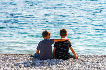 Two boys sitting on the beach, talking and throwing rocks into the sea