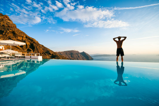 Silhouette of unrecognizable man standing on the edge of an infinity pool looking out over a dramatic Mediterranean view of the Santorini caldera, Greece