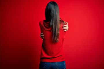Young beautiful brunette woman wearing casual sweater over isolated red background Hugging oneself...