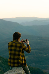 Young boy on a rock taking photos on the mountain with his smartphone, in front of an amazing landscape with sunset. In the middle of nature, with a modern style and casual street clothes. 
