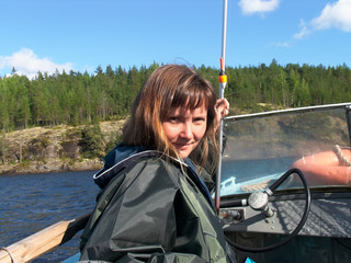 A young woman of Slavic appearance sits on a boat and looks at the center of the frame. In her left hand she holds a fishing rod. In the background you can see the shore of the island and the water.