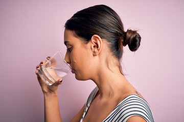 Young beautiful brunette woman drinking glass of healthy water to refreshment standing over isolated pink background