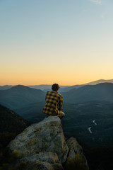 Young boy sitting on rocks watching a mountain landscape, with an incredible sunset, warm sky with...