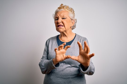Senior beautiful grey-haired woman wearing golden queen crown over white background disgusted expression, displeased and fearful doing disgust face because aversion reaction. With hands raised