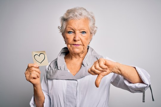 Senior beautiful grey-haired woman holding reminder paper heart over white background with angry face, negative sign showing dislike with thumbs down, rejection concept
