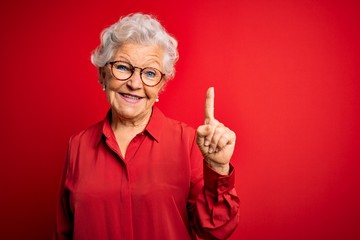 Senior beautiful grey-haired woman wearing casual shirt and glasses over red background showing and...