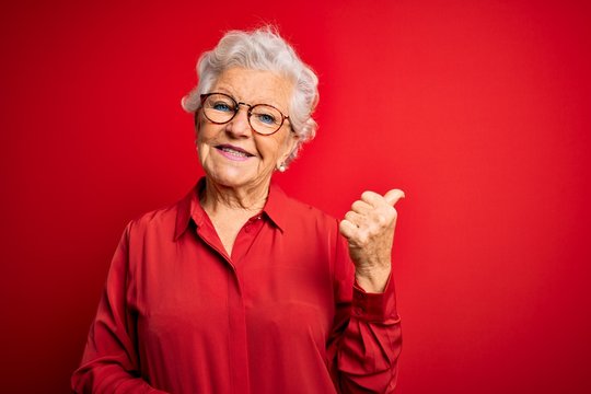 Senior beautiful grey-haired woman wearing casual shirt and glasses over red background smiling with happy face looking and pointing to the side with thumb up.