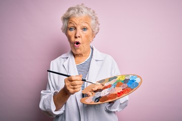 Senior beautiful grey-haired artist woman painting using brush and palette over pink background...