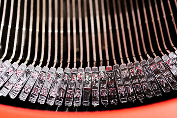 A close-up or a macro shot of the typebars of a retro-style manual typewriter encased in a hinged red top cover.