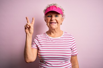 Obraz na płótnie Canvas Senior beautiful sporty woman wearing sport cap standing over isolated pink background smiling with happy face winking at the camera doing victory sign. Number two.