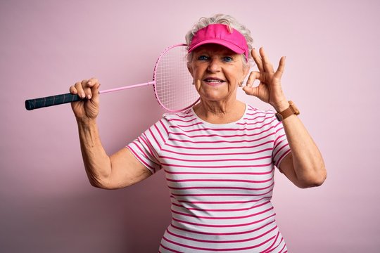 Senior beautiful sportswoman holding badminton racket over isolated pink background doing ok sign with fingers, excellent symbol - Powered by Adobe