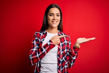 Young beautiful brunette woman wearing casual shirt standing over isolated red background amazed and smiling to the camera while presenting with hand and pointing with finger.