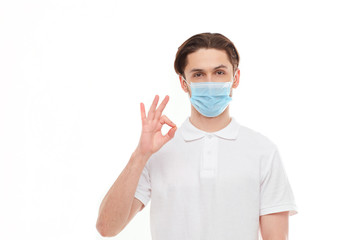 A man in a white T-shirt of European appearance in a medical mask, shows a thumb up. In the mask you are protected from covid 19 viruses.
