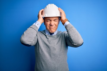 Middle age handsome grey-haired engineer man wearing safety helmet over blue background suffering from headache desperate and stressed because pain and migraine. Hands on head.