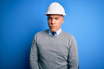 Middle age handsome grey-haired engineer man wearing safety helmet over blue background skeptic and...