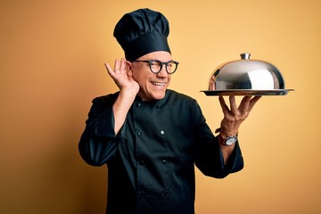 Middle age handsome grey-haired waiter man wearing cooker uniform and hat holding tray smiling with...