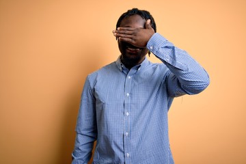Fototapeta premium Young handsome african american man wearing shirt and glasses over yellow background smiling and laughing with hand on face covering eyes for surprise. Blind concept.
