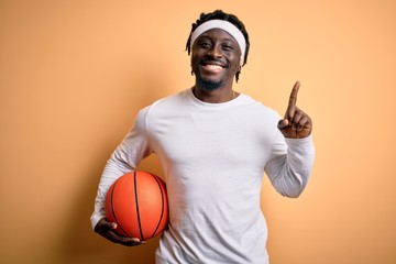 Young african american sportsman doing sport holding basketball ball over yellow background...