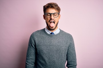 Young handsome man with beard wearing glasses and sweater standing over pink background sticking tongue out happy with funny expression. Emotion concept.