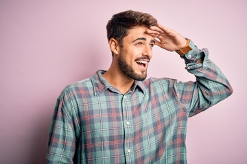 Young handsome man with beard wearing casual shirt standing over pink background very happy and smiling looking far away with hand over head. Searching concept.