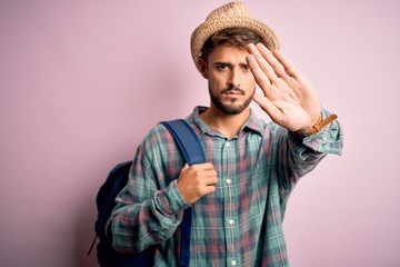 Young tourist man on vacation wearing backpack and summer hat over pink background with open hand doing stop sign with serious and confident expression, defense gesture