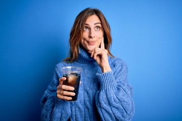 Young beautiful brunette woman drinking glass with cola refreshment over blue background serious...
