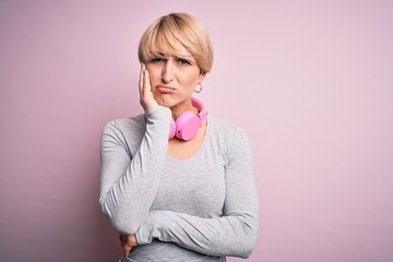 Young blonde woman with short hair wearing headphones on neck over pink background thinking looking tired and bored with depression problems with crossed arms.