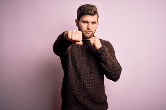 Young blond man with beard and blue eyes wearing casual sweater over pink background Punching fist to fight, aggressive and angry attack, threat and violence