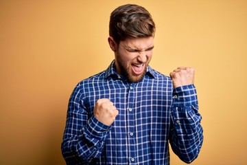 Young blond businessman with beard and blue eyes wearing shirt over yellow background celebrating surprised and amazed for success with arms raised and eyes closed. Winner concept.