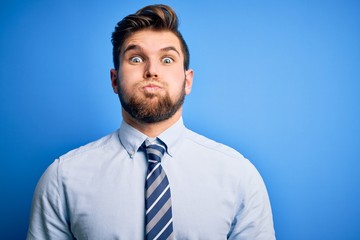 Young blond businessman with beard and blue eyes wearing elegant shirt and tie standing puffing cheeks with funny face. Mouth inflated with air, crazy expression.