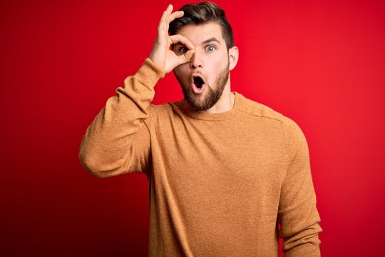 Young blond man with beard and blue eyes wearing casual sweater over red background doing ok gesture shocked with surprised face, eye looking through fingers. Unbelieving expression.