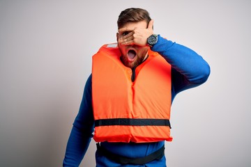 Young blond tourist man with beard and blue eyes wearing lifejacket over white background peeking...