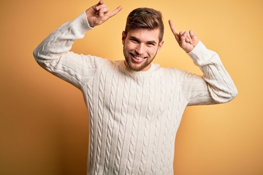 Young blond man with beard and blue eyes wearing white sweater over yellow background smiling amazed and surprised and pointing up with fingers and raised arms.